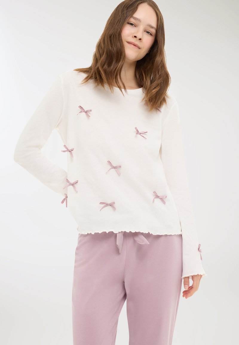 Giovane donna che indossa una maglietta bianca a maniche lunghe con piccoli fiocchi rosa abbinata a pantaloni viola chiaro con cordoncino su uno sfondo neutro.