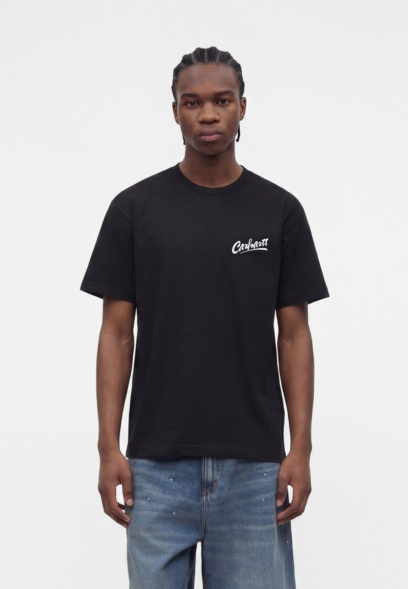 Ung mand med flettet hår iført en sort Carhartt T-shirt og blå bukser sprøjtet med maling, står mod en ensfarvet hvid baggrund.