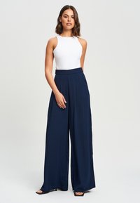 Bijela bezrukava bodi kombiniran s visokim, širokim hlačama u mornarskoj plavoj boji. Glatka tkanina s plisiranjem; model nosi crne sandale na visoku petu.