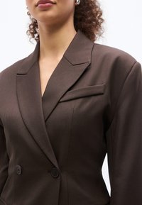 Γυναίκα που φοράει ένα σκούρο καφέ ραμμένο διπλόμπουμπο σακάκι με γιακά notch και δύο κουμπιά, εμφανιζόμενη από τους ώμους έως το μέσο του κορμού.