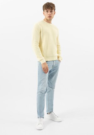 Mladý muž stojící, oblečený v světle žlutém svetru, světle modrých džínách a bílých teniskách, na jednobarevném bílém pozadí.