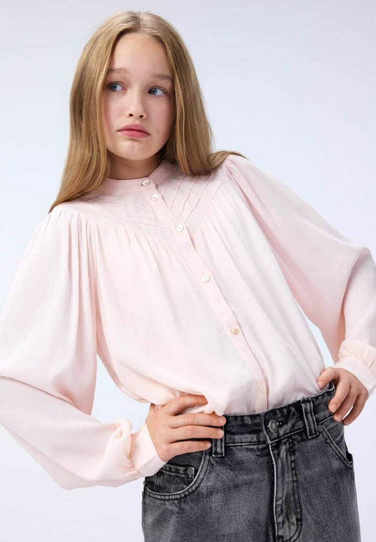 Ljusrosa blus med långa, puffiga ärmar, knapplås framtill och dekorativa veck. Kombineras med höga, mörkgrå denimjeans.