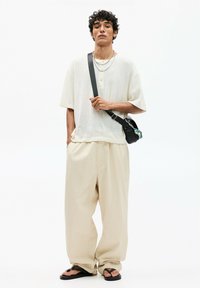 Beige loose-fitting trousers paired with a light, textured short-sleeve shirt. Accessorized with a black crossbody bag and flip-flops.