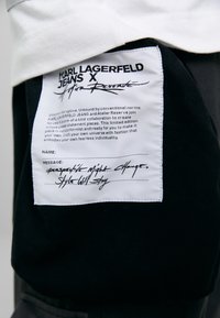 Zwarte stoffen label met witte tekst en handgeschreven accenten. Bevat merknaam, een boodschap over individualiteit en verzorgingsinstructies.
