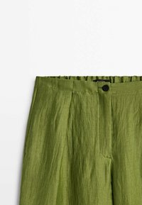 High-waisted olive green linen trousers with pleats, a black button closure, and an elastic waistband at the back.