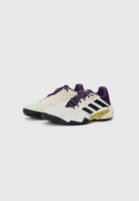 Chaussures de sport avec un dessus en mesh blanc, des accents violets, des rayures noires et une contrepartie de talon dorée. Semelle extérieure en caoutchouc noir texturé pour une bonne adhérence.