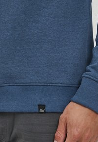 Kék pulóver textúrális anyagból, bordázott mandzsettával és szegéllyel, alján egy kis fekete léccel. Egyszerű, modern dizájn.