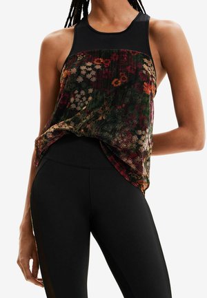 Femme portant un haut noir sans manches avec un panneau transparent à motif floral et un legging noir ajusté, debout avec un bras derrière le dos.