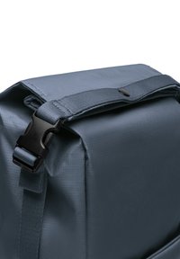 Sac à dos en nylon gris avec un rabat supérieur, équipé d'une boucle en plastique noir et d'un tissu texturé. Dispose d'une poche latérale pour le stockage.