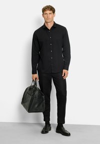 Mann in schwarzem Hemd und Hose, der eine schwarze Lederreisetasche hält, steht vor einem einfarbigen, hellen Hintergrund.