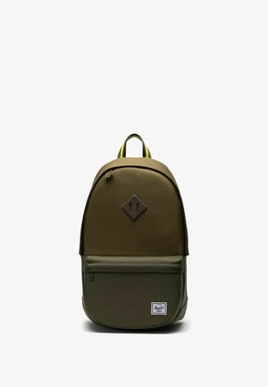 Sac à dos en tissu vert olive avec une forme arrondie, poche avant à fermeture éclair, écusson noir en forme de losange, et poignée supérieure rayée jaune.