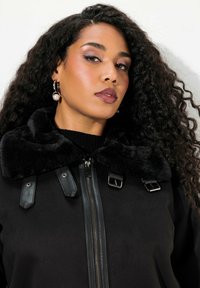 Donna con lunghi capelli ricci indossa una giacca nera con colletto in pelliccia sintetica, orecchini a cerchio d'argento con perle e un trucco sobrio.