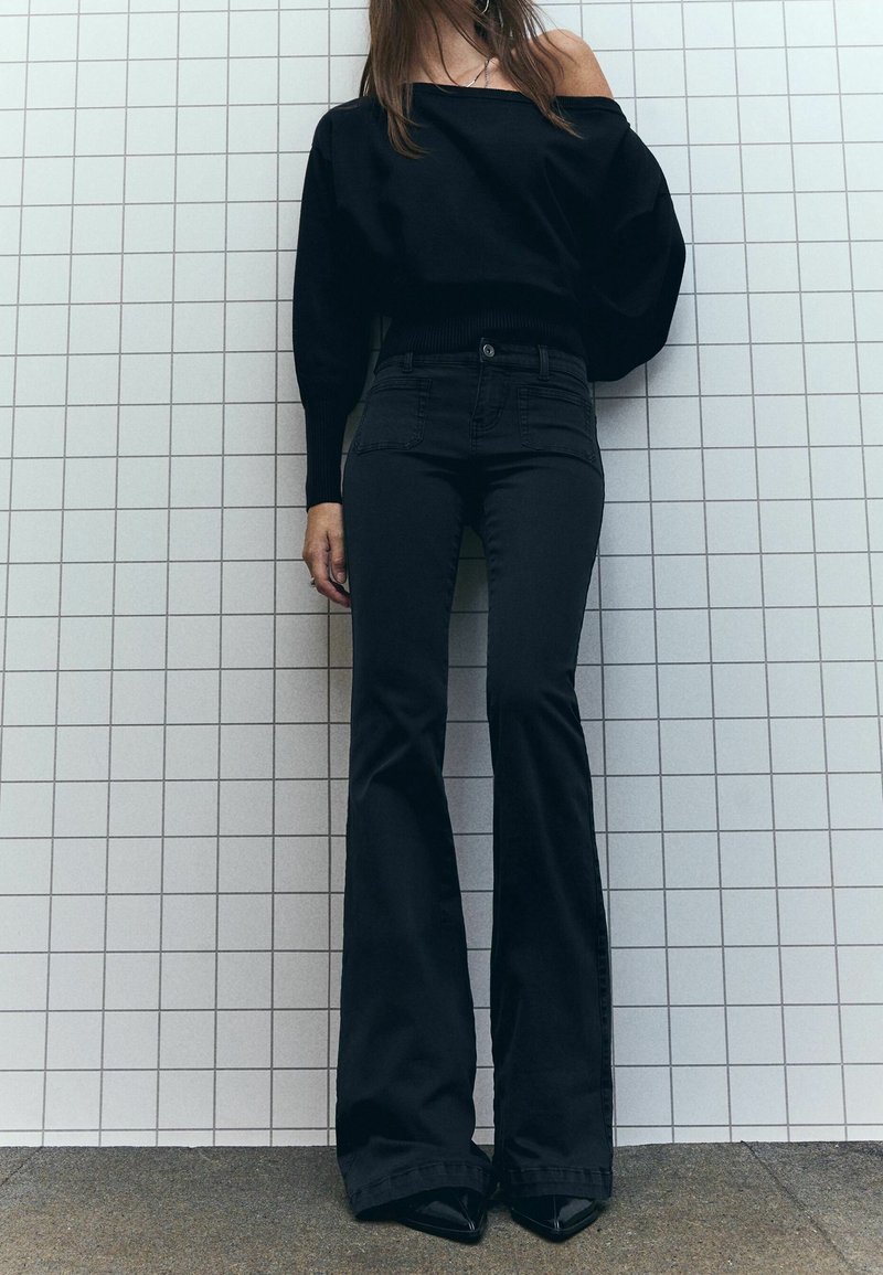 Femme debout contre un mur carrelé blanc, portant un pull noir épaules dénudées, un pantalon noir évasé taille haute et des chaussures noires pointues.