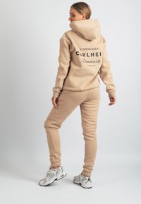 Beige väritetty huppari, jossa on logopainatus takana, yhdistetty samansävyisiin collegehousuihin. Molemmat on valmistettu pehmeästä kankaasta ja niissä on resorit hihansuissa.