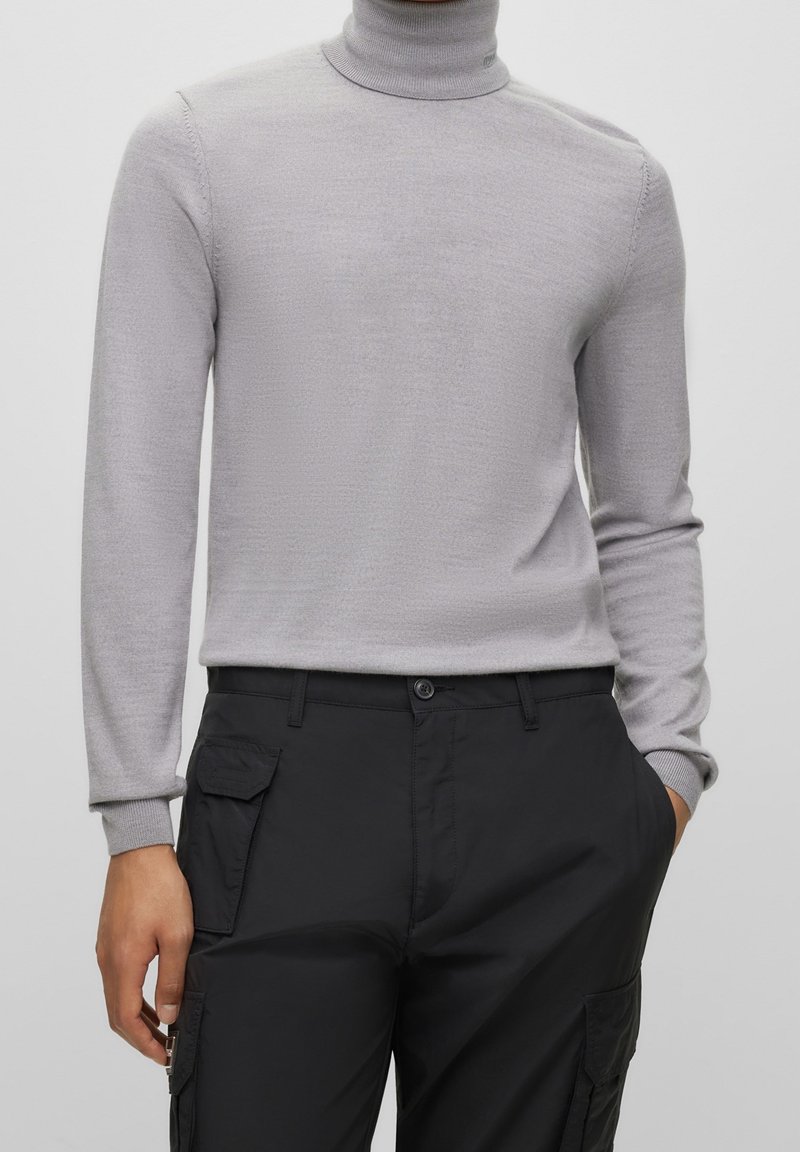 Helles grauer Rollkragenpullover mit langen Ärmeln und strukturiertem Strick. Kombiniert mit schwarzen Cargo-Hosen mit Seitentaschen und Knopfverschluss.