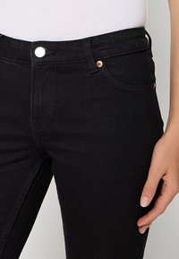 Gros plan d'une personne portant un jean noir moulant avec des rivets en cuivre, un bouton argenté et un haut blanc, la main reposant sur le côté.