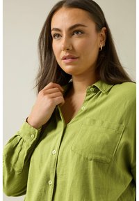 Camisa de lino verde claro con botones y mangas largas, un bolsillo en el pecho y botones redondos. La tela tiene un acabado texturizado.