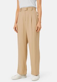 Beige wijde broek met plooien aan de voorkant, een hoge taille en een knoopsluiting, gecombineerd met witte sneakers. Soepele textuur.