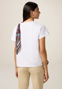 Tricou alb cu mânecă scurtă, cu o eșarfă cu model maroniu atașată la spate, asortat cu pantaloni bej și o brățară din aur.