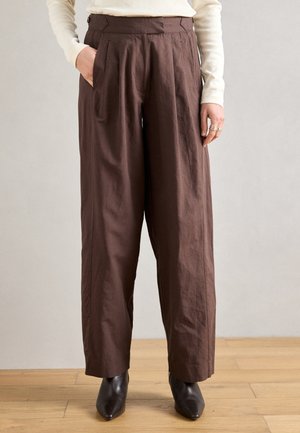 Brązowe spodnie z szerokimi nogawkami, z plisami, bocznymi kieszeniami i gładką fakturą, zestawione z dopasowanymi czarnymi butami z zaostrzonym noskiem.
