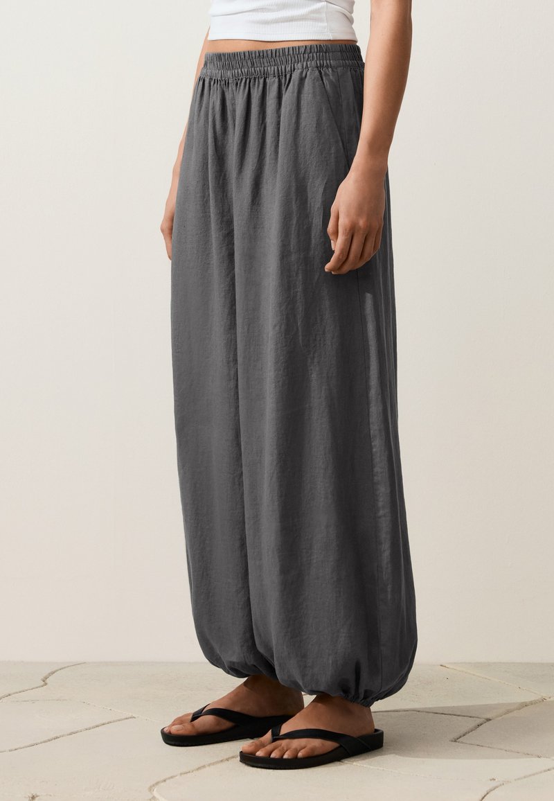 Personne portant un pantalon ample gris foncé avec poignets et taille élastiqués, un haut court blanc et des tongs noires, debout sur un sol carrelé.