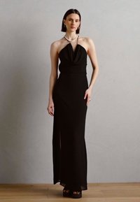 Rochie neagră cu guler halter, cu un decolteu în formă de cowl, talie ajustată și fante laterale. Textură de țesătură netedă, design minimalist, completată cu toc platform negru.
