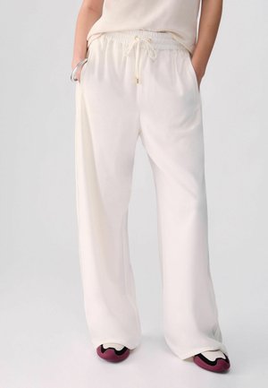 Personne portant un pantalon blanc ample à cordon, un haut crème, les mains dans les poches, un bracelet en argent et des baskets blanches avec des touches roses et noires.