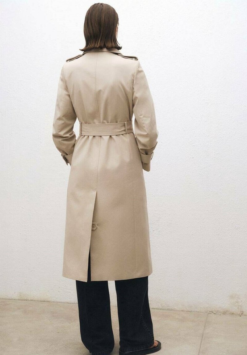 Trench-coat beige avec une taille ceinturée, des épaulettes et une fente à l'arrière. Associé à un jean foncé et des sandales.