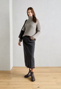 Pull à col roulé en tricot côtelé gris, jupe midi en denim noir avec ourlet effiloché, et bottes cheville en cuir marron. Sac noir porté sur l'épaule.