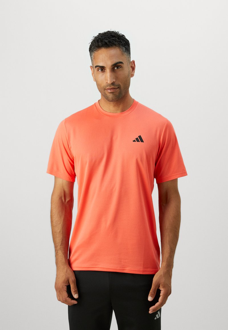 Tricou sportiv coral cu mânecă scurtă, realizat dintr-un material respirabil, având un decolteu rotund și un logo negru pe pieptul stâng.