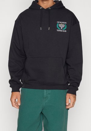 Homme portant un sweat à capuche noir avec le logo "On Vacation Tennis Club" et des raquettes croisées, associé à un pantalon vert, debout devant un fond blanc.