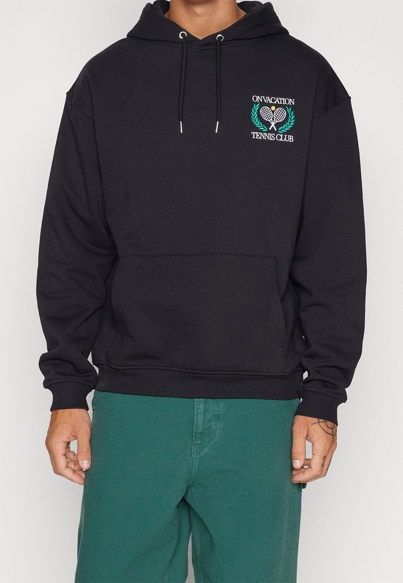 Homme portant un sweat à capuche noir avec le logo "On Vacation Tennis Club" et des raquettes croisées, associé à un pantalon vert, debout devant un fond blanc.