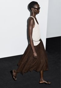 Lichtbeige tanktop, bruine vloeiende rok met een gladde textuur en zwarte teenslippers. Model draagt een donkere zonnebril. Effen achtergrond.