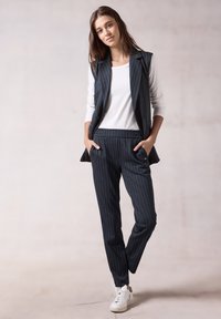 Dunkelblau gestreifter Weste und passende Hose, aus einem weichen Stoff gefertigt. Darunter ein weißes Langarmshirt, kombiniert mit weißen Sneakers.