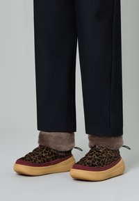 Scarpe con stampa leopardata e una cintura in pelliccia sintetica marrone, dettagli rosso scuro e suola spessa beige. I pantaloni sono neri e leggermente larghi.