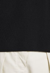 Gros plan sur un pull noir en maille côtelée rentré dans un pantalon plissé blanc cassé.