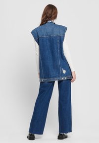 Denimväst i ljusblått med breda ärmhål, råa kanter och sömmar bak. Kombinerad med löst sittande blå jeans och svarta platåskor.