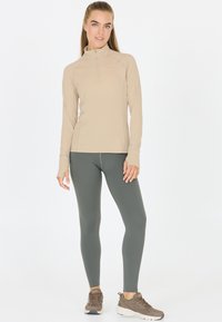 Beige langekleurig top met een ritssluiting kraag, gecombineerd met donker groene leggings. De outfit heeft een getailleerd ontwerp en lichte stoffen.