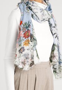 Femme portant un haut blanc à manches longues, un pantalon beige taille haute plissé, et un foulard bleu clair à motif floral enroulé autour du cou.