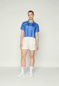 Top cropped listrado azul e roxo com colarinho amarelo, logotipo no peito, combinado com calções brancos. Meias e sapatos brancos completam o look.