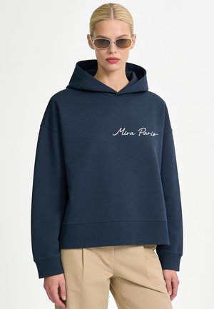 Blond meisje met bruine zonnebril, een marineblauwe hoodie met de tekst ""Mira Paris"" en beige broek tegen een effen witte achtergrond.