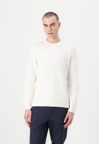 White boucle knit sweater with long sleeves and round neckline, worn with dark blue pants. Smooth texture and casual fit.