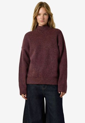 Femme aux cheveux blonds portant un pantalon ample bleu foncé et un pull violet à col montant côtelé, debout et faisant face.