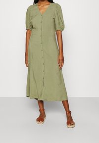Olivgrünes Midi-Kleid, mit Knopfleiste vorne, V-Ausschnitt, Puffärmeln, leichtem Stoff und einem schwingenden Rockdesign. Kombiniert mit braunen Sandalen.