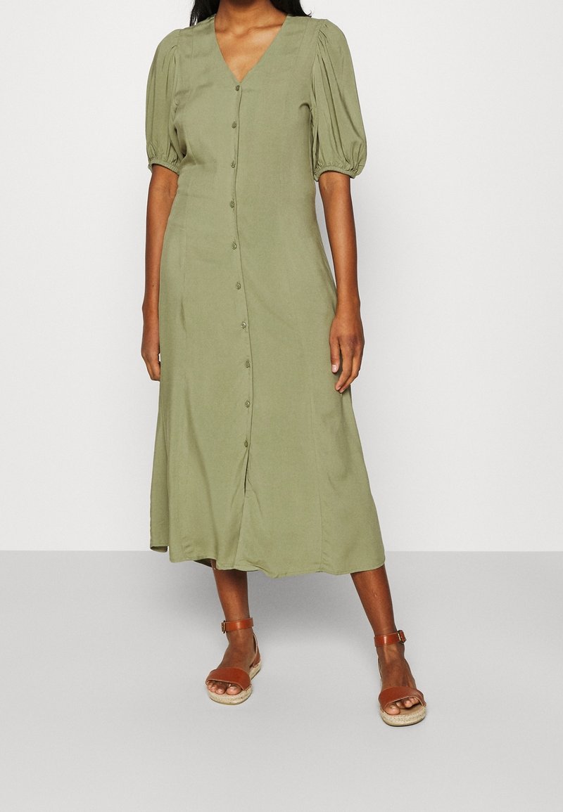 Olivgrünes Midi-Kleid, mit Knopfleiste vorne, V-Ausschnitt, Puffärmeln, leichtem Stoff und einem schwingenden Rockdesign. Kombiniert mit braunen Sandalen.