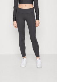 Kvinna i svarta ribbade leggings med sidoficka och vita Nike-sneakers, stående mot en enkel ljus bakgrund.