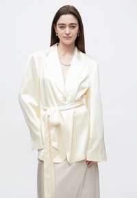  Dramblio kaulo spalvos satino švarkas su diržo juosmeniu, plačiomis atlapais ir dviem kišenėmis; derinamas su kreminės spalvos suknia. Lygus tekstūros, laisvo silueto modelis.