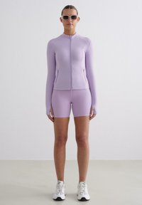 Svetle fialový sportovní outfit s dlouhým zipovým sakem a přiléhavými šortkami. Hladká tkanina s elegantním designem. V kombinaci s bílými teniskami.