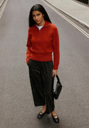 Donna in piedi su una strada che indossa un maglione rosso con cerniera, pantaloni neri larghi con motivo, ballerine nere, tiene in mano una borsa nera.