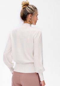 Kremowy sweter o luźnym kroju z długimi, bufiastymi rękawami i żebrowanym wykończeniem. Lekki materiał o gładkiej teksturze i wysokim kołnierzu.